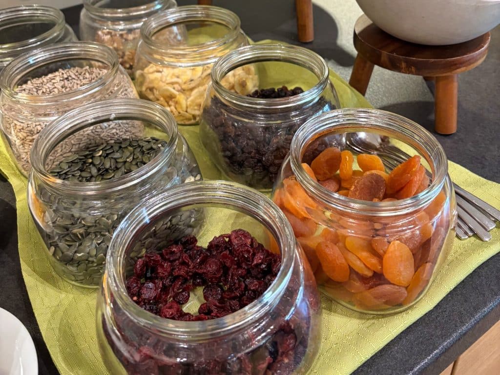 Mehrere offene Glasbehälter auf einem Frühstücksbuffet im Susato Hotel, gefüllt mit verschiedenen Toppings wie getrockneten Aprikosen, Cranberrys, Rosinen, Bananenchips, Kürbiskernen und Sonnenblumenkernen.