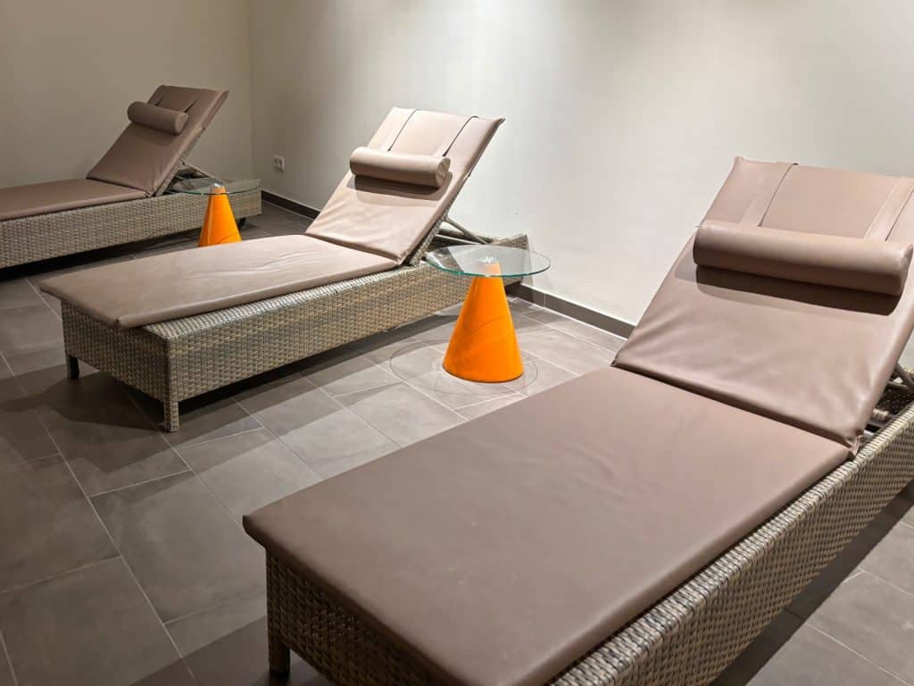 Drei beigefarbene Rattan-Liegen mit passenden Polstern und Nackenrollen stehen im Ruhebereich der Sauna des Susato Hotels neben kleinen Glastischen mit auffälligen orangefarbenen Kegelfüßen.