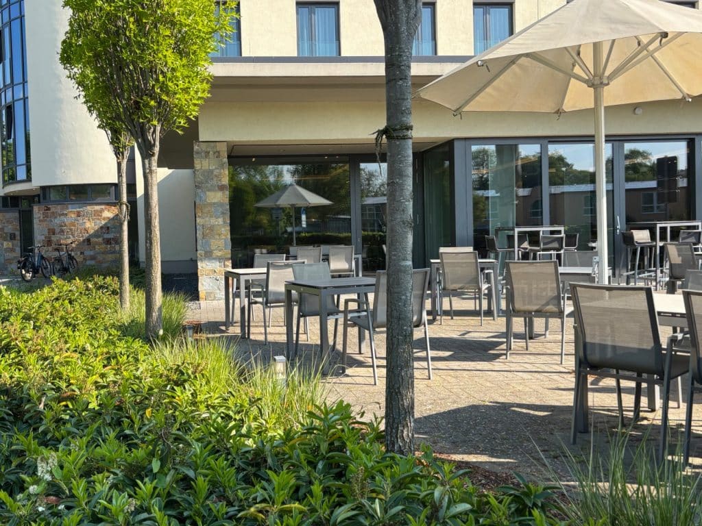 Die sonnige Terrasse des Hotel in Soest ist mit modernen grauen Tischen und Stühlen unter großen Sonnenschirmen ausgestattet, umgeben von gepflegten Grünanlagen und jungen Bäumen.