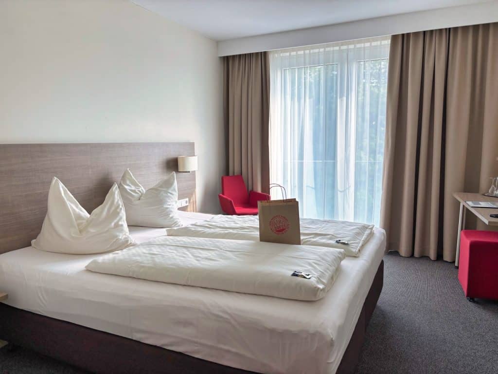 Ein helles Hotelzimmer in Soest mit einem gemachten Doppelbett, auf dem eine braune Papiertüte steht, sowie einem roten Sessel vor einem großen Fenster mit beigen Vorhängen.