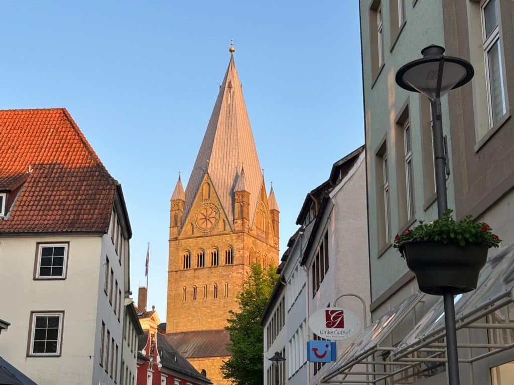 Blick durch eine schmale Gasse in Soest auf den markanten, sandsteinfarbenen Turm der St.-Patrokli-Kirche unter einem klaren blauen Himmel.