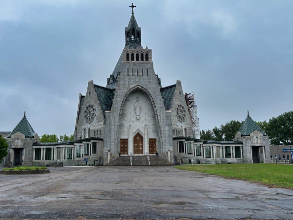 Die monumentale Kirche Notre-Dame-du-Cap in Trois-Rivières aus grauem Stein präsentiert sich mit einer markanten, spitz zulaufenden Fassade, einem zentralen Turm mit Kreuz und zwei symmetrischen Seitenflügeln unter einem bewölkten Himmel. Vor dem imposanten Sakralbau erstreckt sich ein weitläufiger, asphaltierter Vorplatz, während im Hintergrund grüne Bäume und ein Teil eines Baugerüsts zu sehen