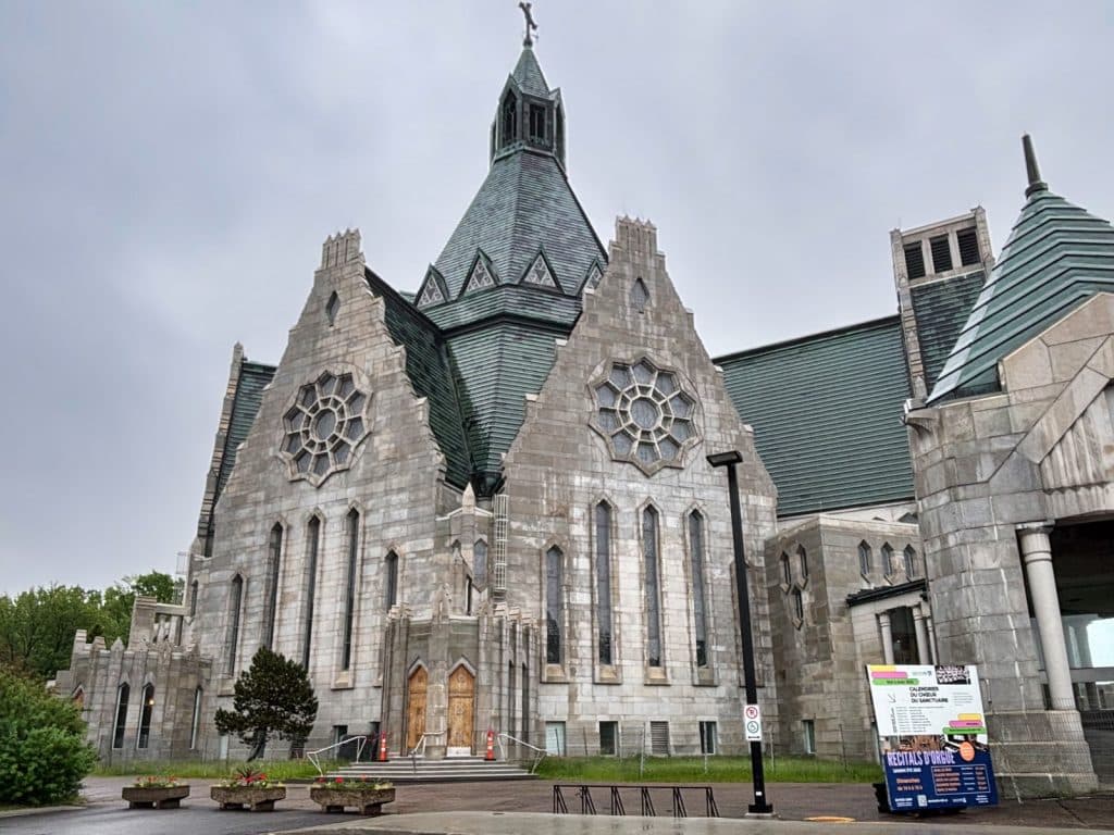 Außenansicht der imposanten, neugotischen Steinkirche Notre-Dame-du-Cap in Trois-Rivières mit markanten Rosettenfenstern und einem hohen, grünlichen Kupferdach unter bewölktem Himmel.