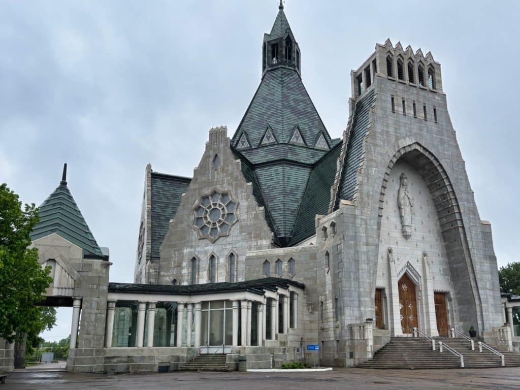Die Außenansicht der monumentalen Kirche Notre-Dame-du-Cap in Trois-Rivières zeigt eine graue Steinfassade mit einem markanten, spitz zulaufenden Kupferdach und einem großen Rundfenster. Rechts führt eine breite Treppe zum Haupteingang, der von einer hohen Statue in einer Nische überragt wird.