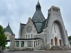 Die Außenansicht der monumentalen Kirche Notre-Dame-du-Cap in Trois-Rivières zeigt eine graue Steinfassade mit einem markanten, spitz zulaufenden Kupferdach und einem großen Rundfenster. Rechts führt eine breite Treppe zum Haupteingang, der von einer hohen Statue in einer Nische überragt wird.