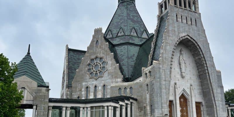Die Außenansicht der monumentalen Kirche Notre-Dame-du-Cap in Trois-Rivières zeigt eine graue Steinfassade mit einem markanten, spitz zulaufenden Kupferdach und einem großen Rundfenster. Rechts führt eine breite Treppe zum Haupteingang, der von einer hohen Statue in einer Nische überragt wird.