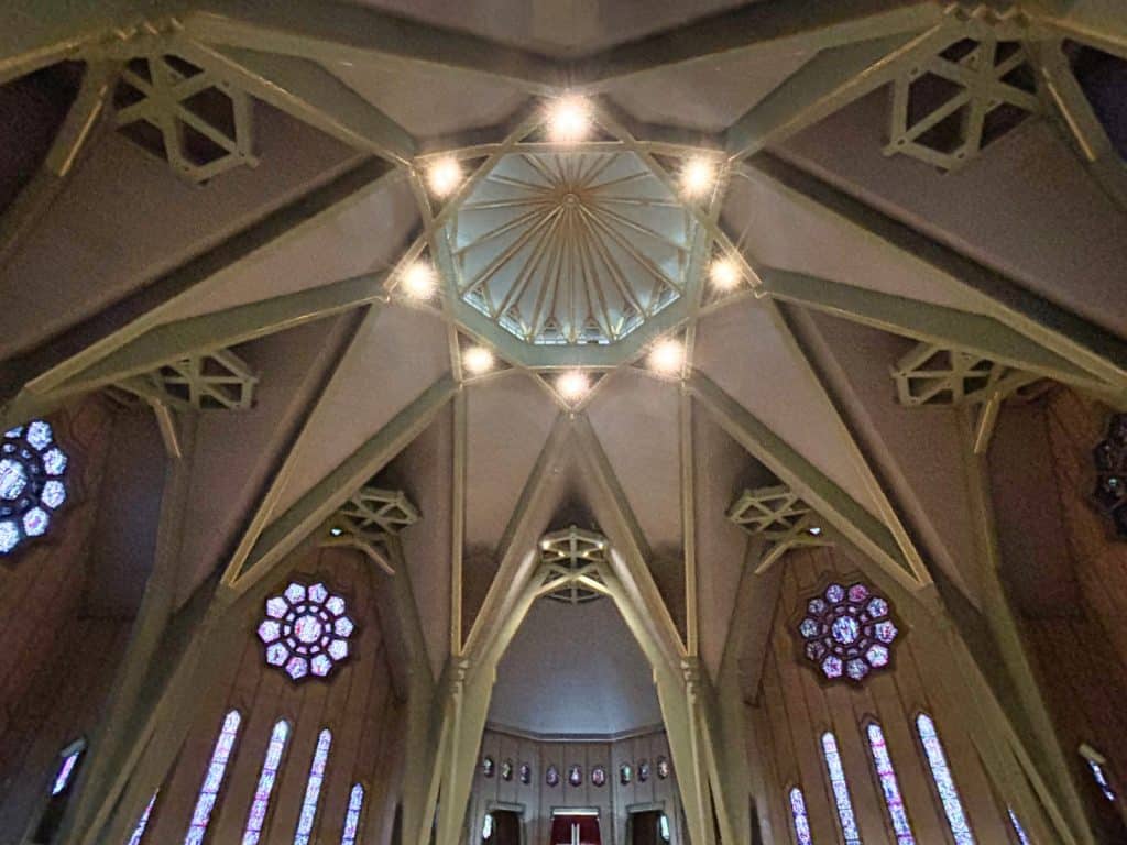 Blick nach oben an die Decke der Kirche Notre-Dame-du-Cap in Trois-Rivières, die durch eine komplexe, sternförmige Betonstruktur mit einem zentralen Oberlicht und farbenfrohen Rosettenfenstern geprägt ist.