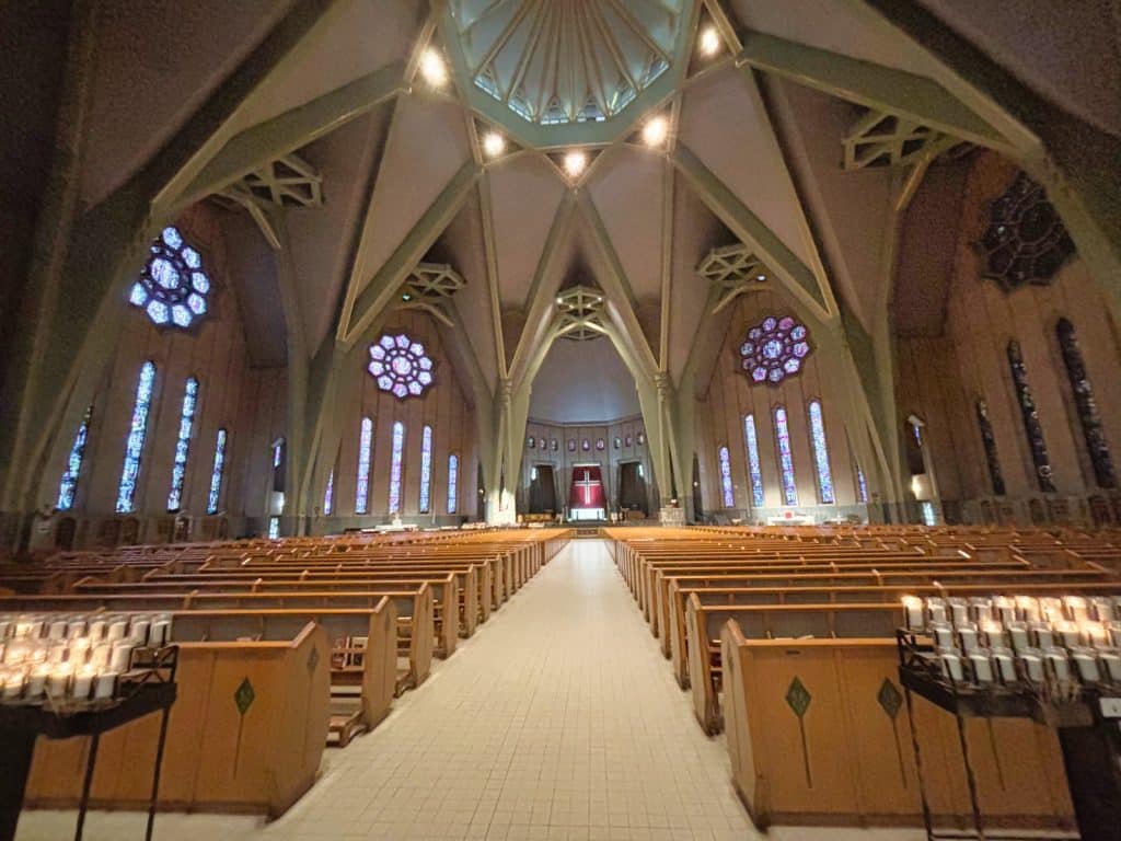 Das weite Innere der Kirche in Trois-Rivières zeigt hohe, fächerförmige Betongewölbe, bunte Rosettenfenster und lange Reihen hölzerner Kirchenbänke, die auf einen fernen Altar mit einem roten Kreuz ausgerichtet sind.