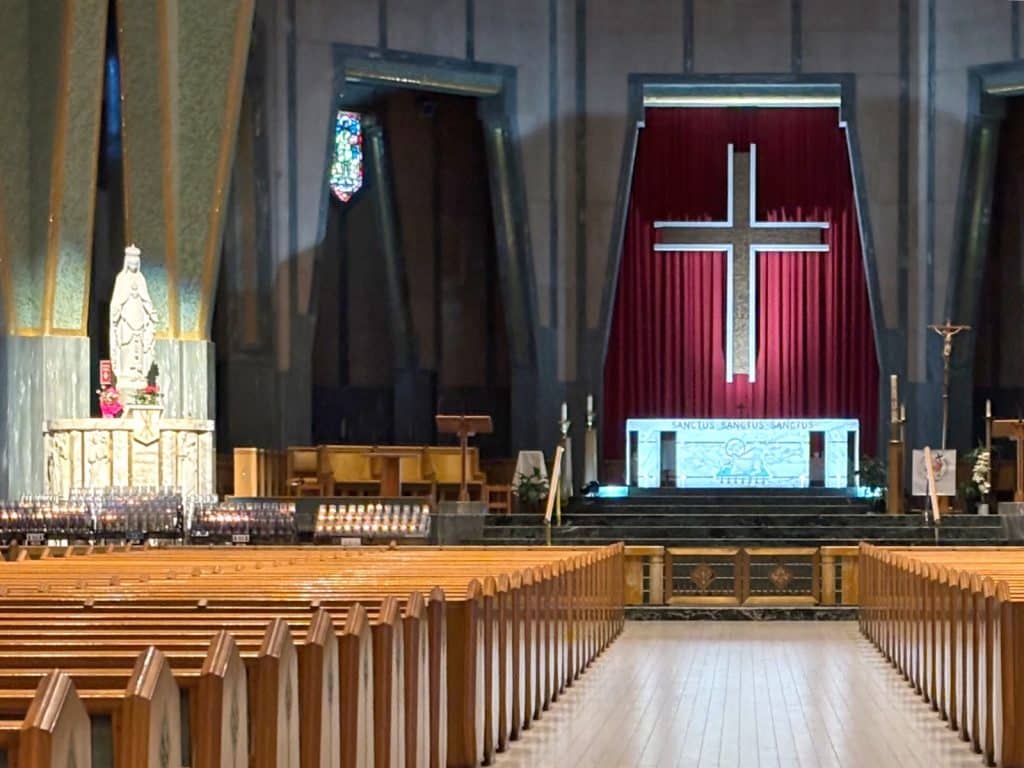 Innenansicht einer Kirche in Trois-Rivières mit hölzernen Kirchenbänken im Vordergrund, einer Marienstatue auf der linken Seite und einem zentralen Altar vor einem großen Kreuz auf rotem Hintergrund.