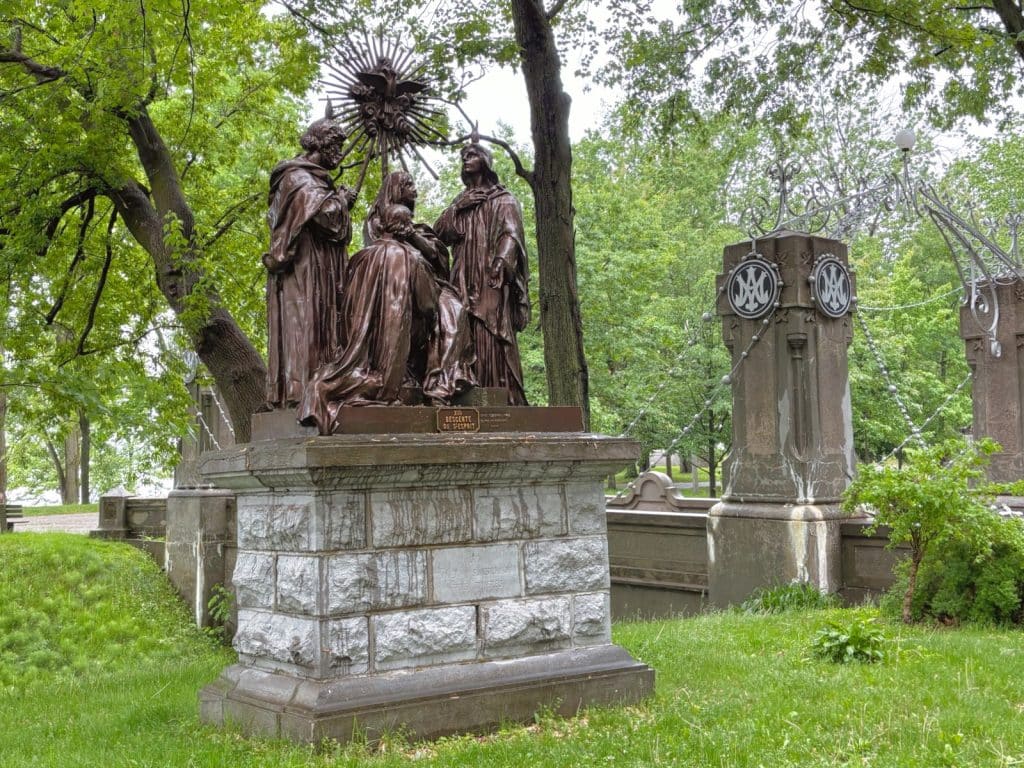 Eine bronzene Figurengruppe auf einem steinernen Sockel stellt eine religiöse Szene dar und ist Teil des Skulpturenwegs an der Kirche in Trois-Rivières, umgeben von grünen Bäumen und einer historischen Steinfassung.