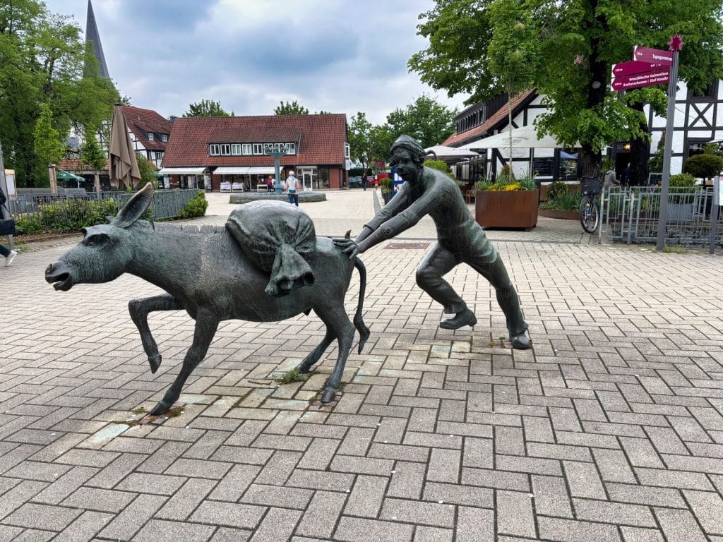 Eine Bronzeskulptur in Bad Sassendorf zeigt einen Mann, der mit vollem Körpereinsatz einen störrischen Esel mit einem schweren Sack auf dem Rücken vor sich herschiebt. Im Hintergrund sind gepflasterte Wege, Fachwerkhäuser und eine herbstliche Parkanlage zu sehen.
