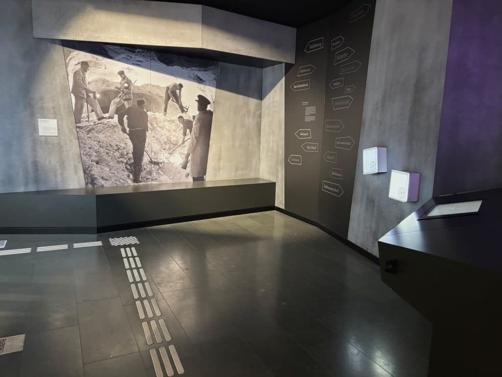 Ein moderner Ausstellungsraum in den Westfälischen Salzwelten in Bad Sassendorf zeigt eine großformatige historische Schwarz-Weiß-Fotografie von Bergleuten bei der Arbeit sowie eine Wand mit zahlreichen Hinweisschildern zu regionalen Orten. Der Raum ist in schlichtem Grau und Schwarz gestaltet, verfügt über taktile Bodenleitlinien für Barrierefreiheit und interaktive Stationen an den Wänden.