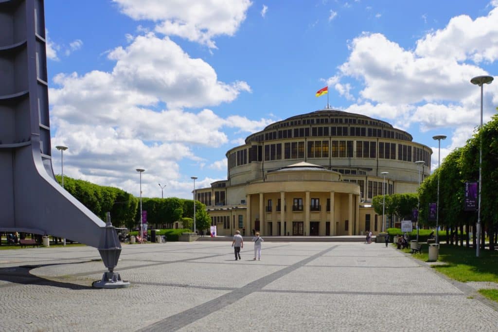 Blick auf die monumentale Front der Jahrhunderthalle in Breslau unter blauem Himmel, davor ein weiter gepflasterter Platz mit Passanten und einer modernen Skulptur am linken Bildrand.