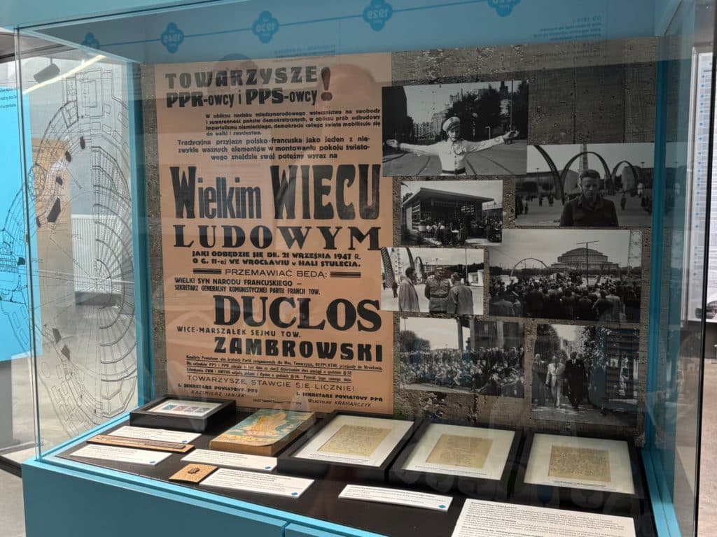 Eine Museumsvitrine im Besucherzentrum der Breslauer Jahrhunderthalle zeigt ein historisches Plakat von 1947 sowie verschiedene Schwarz-Weiß-Fotografien und Dokumente zur Geschichte des Bauwerks.
