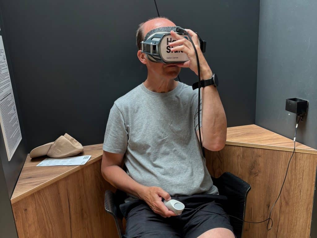 Ein Mann sitzt im Besucherzentrum der Breslauer Jahrhunderthalle und nutzt eine VR-Brille sowie einen Handcontroller für ein virtuelles Erlebnis. Er trägt ein graues T-Shirt und hält das Headset mit einer Hand fest, während er sich in einer hölzernen Sitznische befindet.