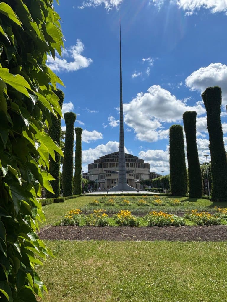 Blick durch grünes Laub auf die Jahrhunderthalle in Breslau, vor der die markante, spitze Stahlnadel Iglica in den blauen Sommerhimmel ragt.