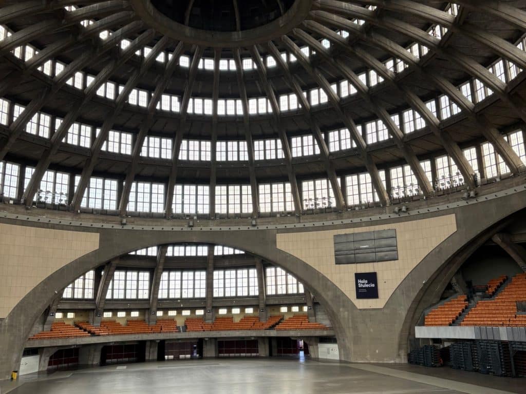 Innenansicht der monumentalen Jahrhunderthalle in Breslau mit ihrer markanten, weitgespannten Betonkuppel, zahlreichen Fensterreihen und den markanten Zuschauerrängen mit orangefarbenen Sitzen.
