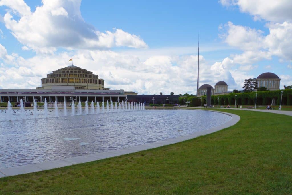 Die Jahrhunderthalle in Breslau mit ihrer markanten Kuppel hinter einer großen Brunnenanlage mit Wasserfontänen, flankiert von einem modernen Nebengebäude und der hohen Iglica-Nadel unter bewölktem Himmel.