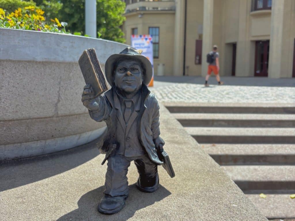 Eine kleine Bronzestatue eines Zwerges im Anzug mit Hut und Buch steht auf einer Mauer vor der Jahrhunderthalle in Breslau.