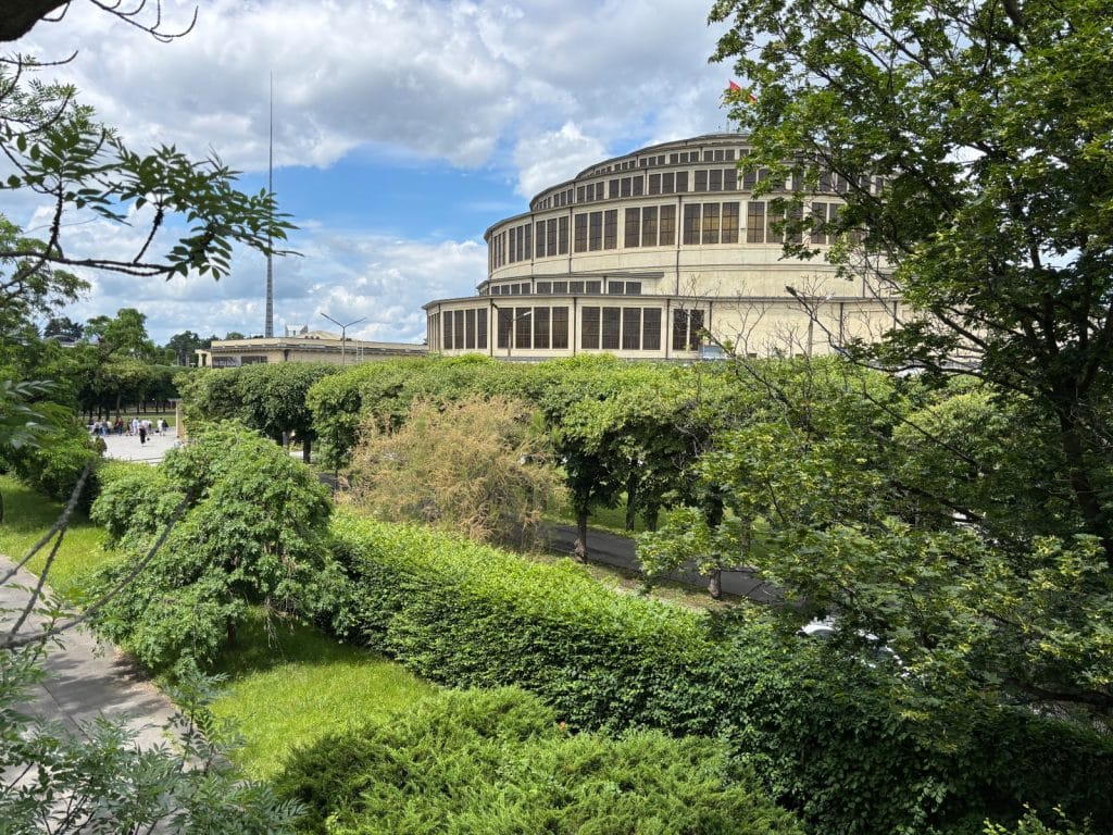 Blick durch üppiges grünes Gebüsch und Bäume auf die monumentale Breslauer Jahrhunderthalle mit ihrer markanten Kuppelarchitektur unter einem leicht bewölkten Himmel. Im Hintergrund ragt links die schlanke Iglica-Nadel empor.
