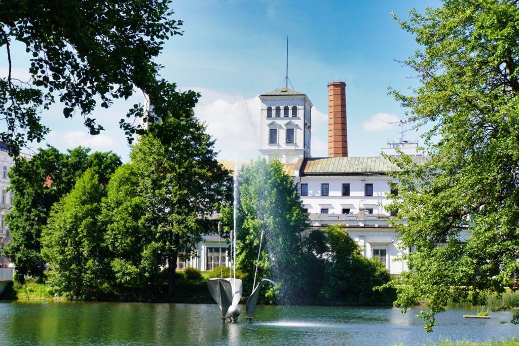 Blick über einen See mit Springbrunnen auf die Weiße Fabrik Łódź, das von grünen Bäumen umgeben ist und einen markanten Ziegelschornstein besitzt.