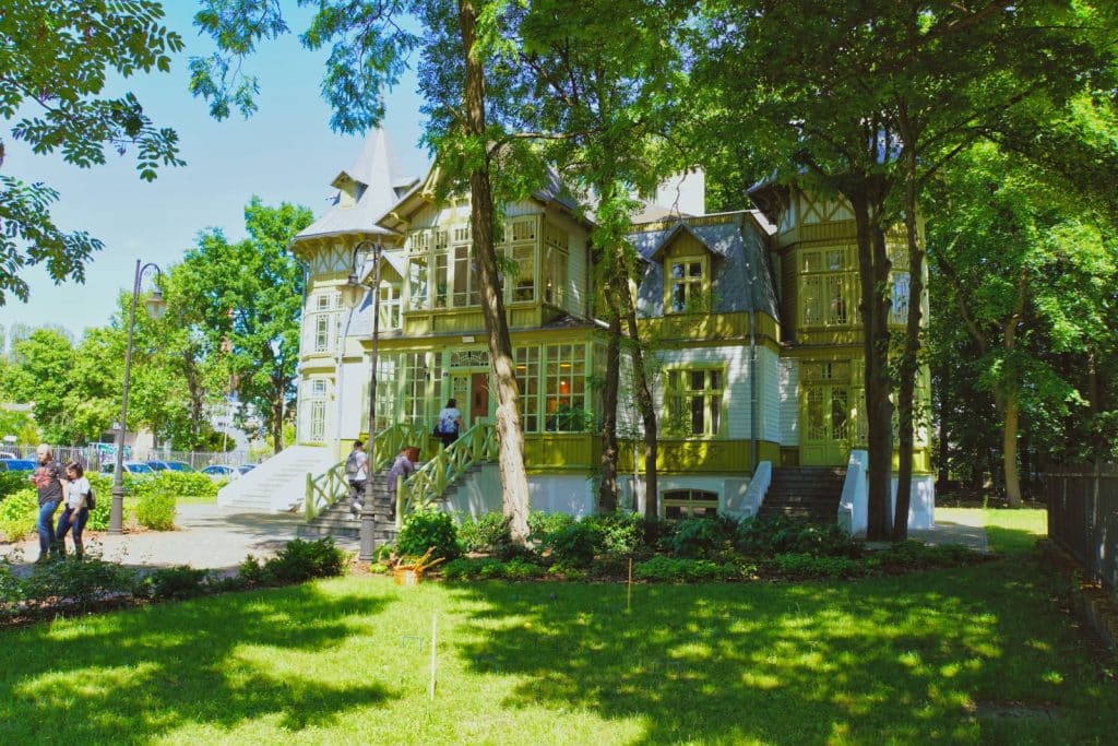 Das hölzerne Sommerhaus bei der Weißen Fabrik Łódźpräsentiert sich als prachtvolle, hellgrüne Villa mit verspielten Erkern und Türmchen, umgeben von hohen, schattenspendenden Bäumen und einer gepflegten Rasenfläche. Mehrere Personen steigen die Treppen zum Eingang hinauf, während im Vordergrund eine sonnige Parkanlage zu sehen ist.