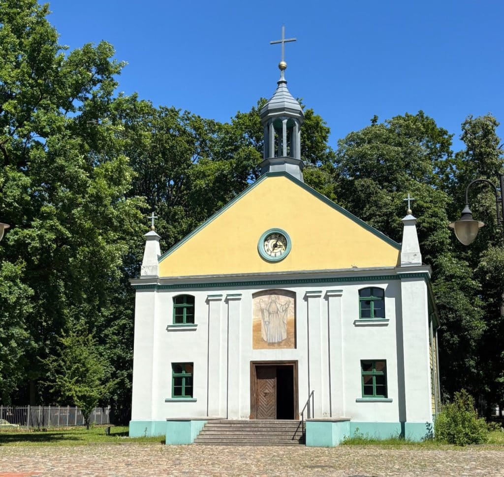 Die kleine, hellgelb und weiß gestrichene Holzkirche im Freilichtmuseum der Lodzer Textilindustrie steht unter blauem Himmel vor grünen Bäumen und zeigt ein Wandbild über dem Eingang sowie ein kleines Türmchen mit Kreuz.