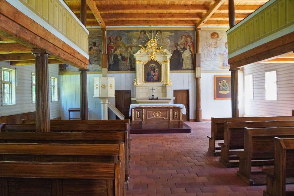 Innenansicht der hölzernen Kirche im Textilmuseum in Łódź mit Blick auf den Altar, Wandmalereien und hölzerne Kirchenbänke unter einer Galerie.