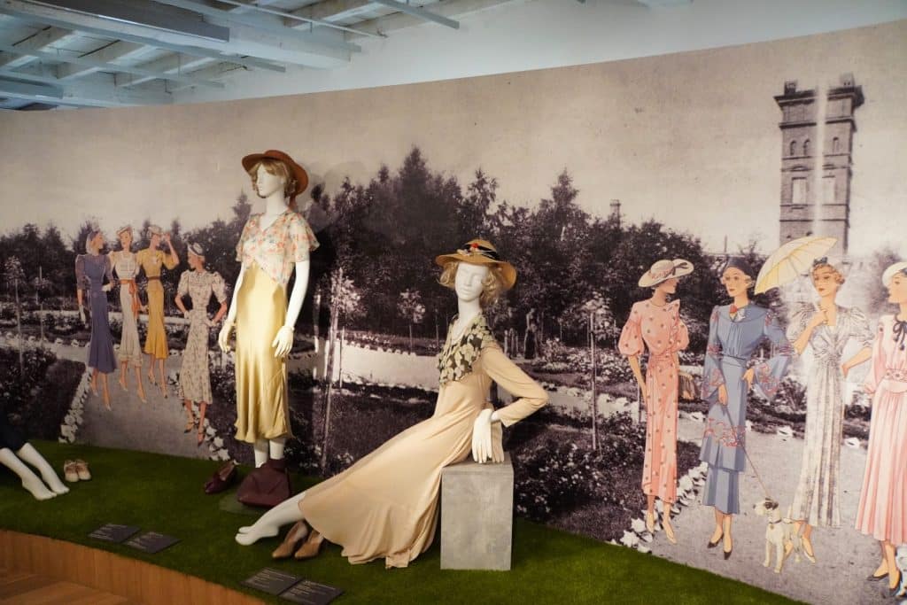 Eine Ausstellung im Textilmuseum in Łódź zeigt Schaufensterpuppen in eleganter Damenmode der 1930er Jahre vor einer großformatigen historischen Fotowand eines Parks.