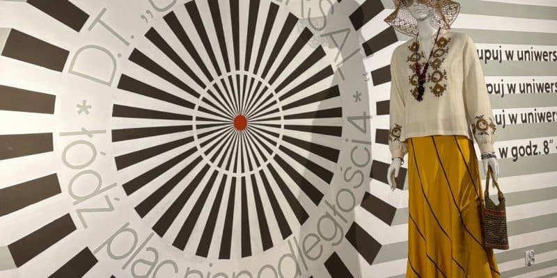 Eine Ausstellung in der Weiße Fabrik Łódź zeigt ein großflächiges, kreisförmiges Grafikdesign an der Wand und eine Schaufensterpuppe in einem modischen Ensemble aus bestickter weißer Bluse, langem gelbem Rock und Strohhut.