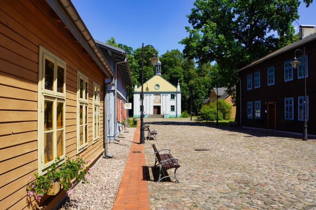 Blick auf eine gepflasterte Straße im Museumsdorf des Zentralen Textilmuseums in Łódź, gesäumt von historischen Holzhäusern und Holzbänken, die zu einer kleinen weißen Kirche im Hintergrund führt.