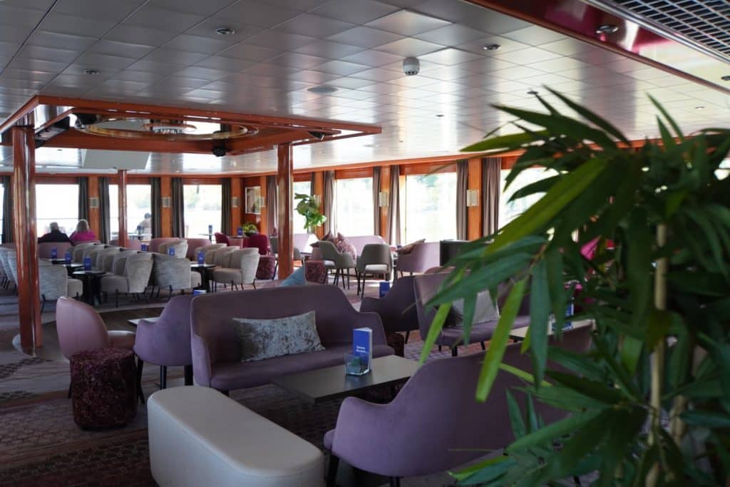 Der Barbereich der Maxima nicko cruises mit violetten Polstersofas, Sesseln und kleinen Tischen, die vor großen Panoramafenstern in einem hellen, modern gestalteten Salon angeordnet sind.
