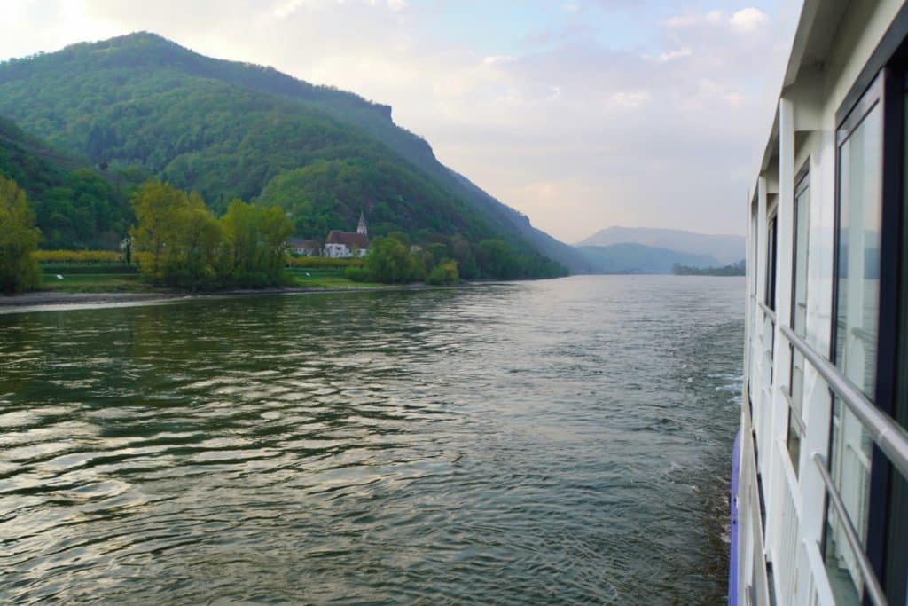 Blick von Bord des Flusskreuzfahrtschiffs Maxima nicko cruises auf die Donau, die an bewaldeten Hügeln und einer kleinen Kirche am Ufer vorbeiführt.