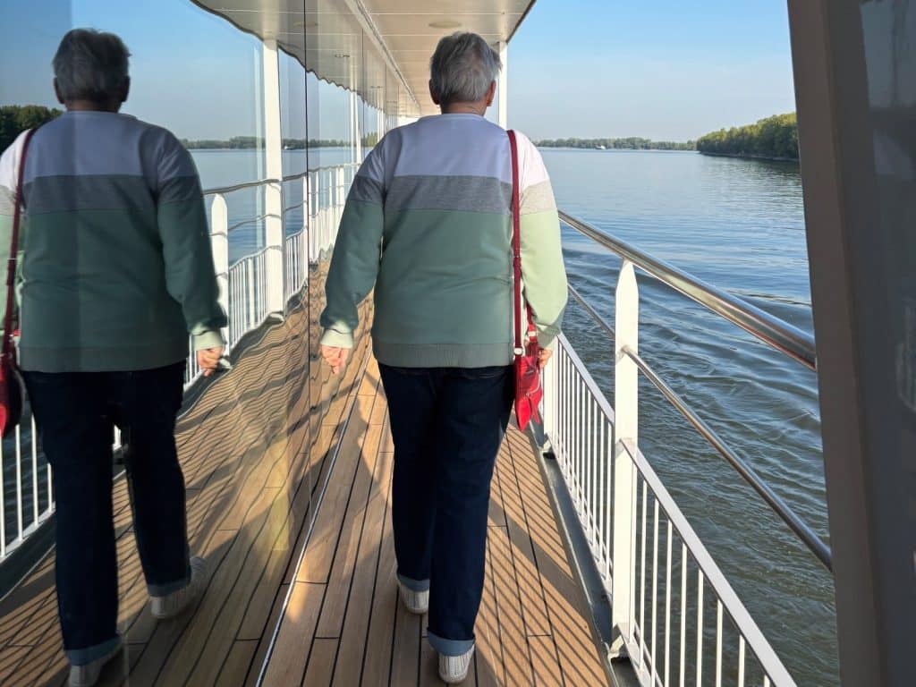 Eine Person geht über das sonnige Außendeck der Maxima, wobei sich ihre Gestalt in der großen Fensterfront der Kabinen spiegelt, während das Schiff an einer bewaldeten Flusslandschaft vorbeifährt.