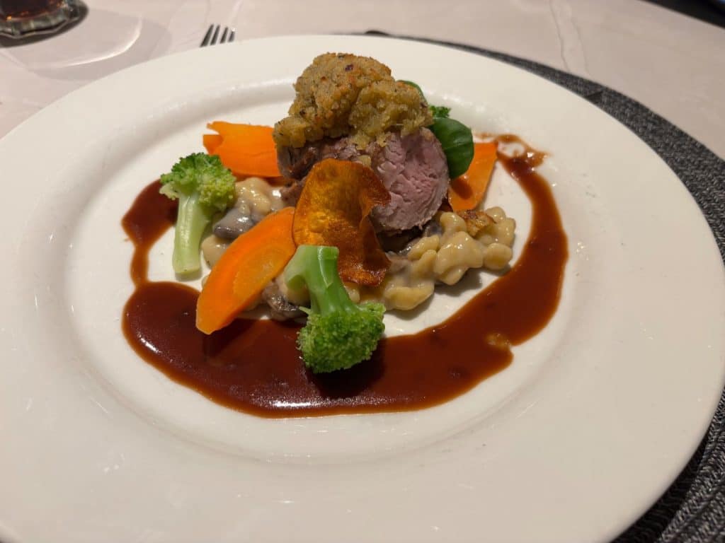 Ein von Johann Lafer kreierter Hauptgang auf der Maxima nicko cruises, bestehend aus einem rosa gebratenen Fleischstück mit Kräuterkruste, serviert auf Spätzle mit Brokkoli, Karotten und einer dunklen Sauce.