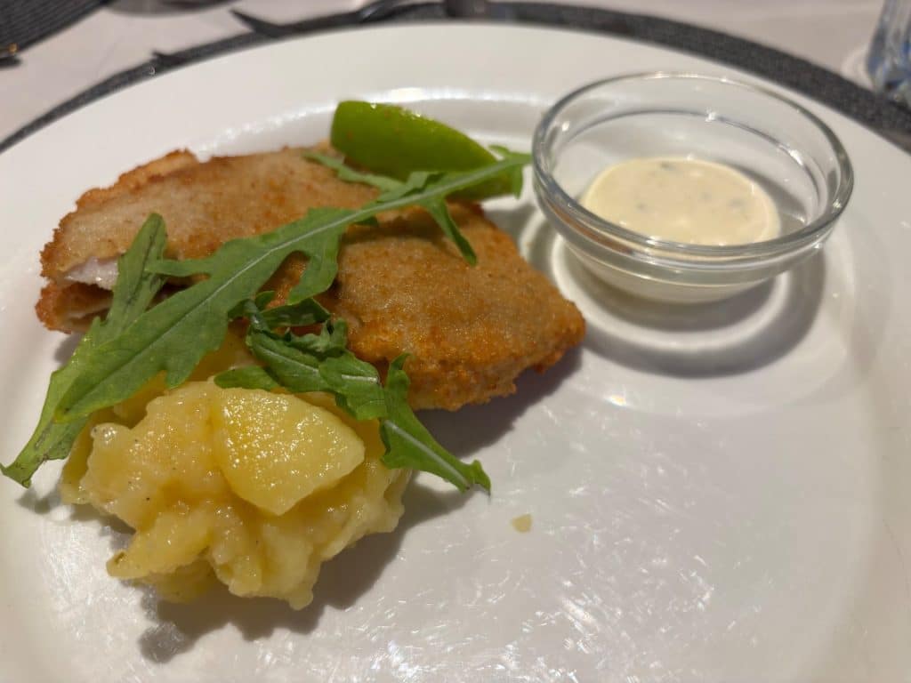 Hauptgang des Piratenessens auf der Maxima: Ein paniertes Fischfilet serviert mit Kartoffelsalat, einer Limettenspalte, Rucola und einer cremigen Sauce in einem kleinen Glasschälchen.