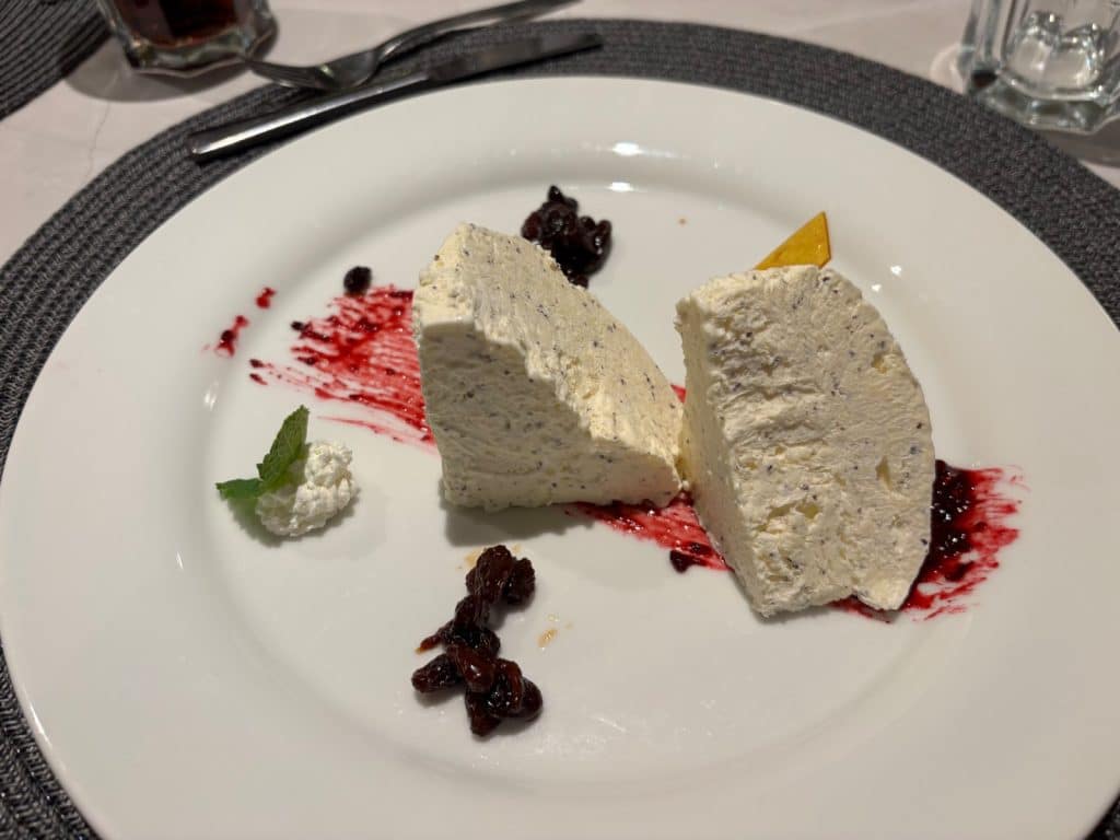 Die Nachspeise des Piratenessens auf der Maxima zeigt zwei cremige, hellbeige Mousse-Keile mit dunklen Sprenkeln auf einem weißen Teller, garniert mit roten Beerensaucen-Strichen, dunklen Beeren, einem Klecks Sahne mit Minzblatt und einem gelben Schokoladendreieck.