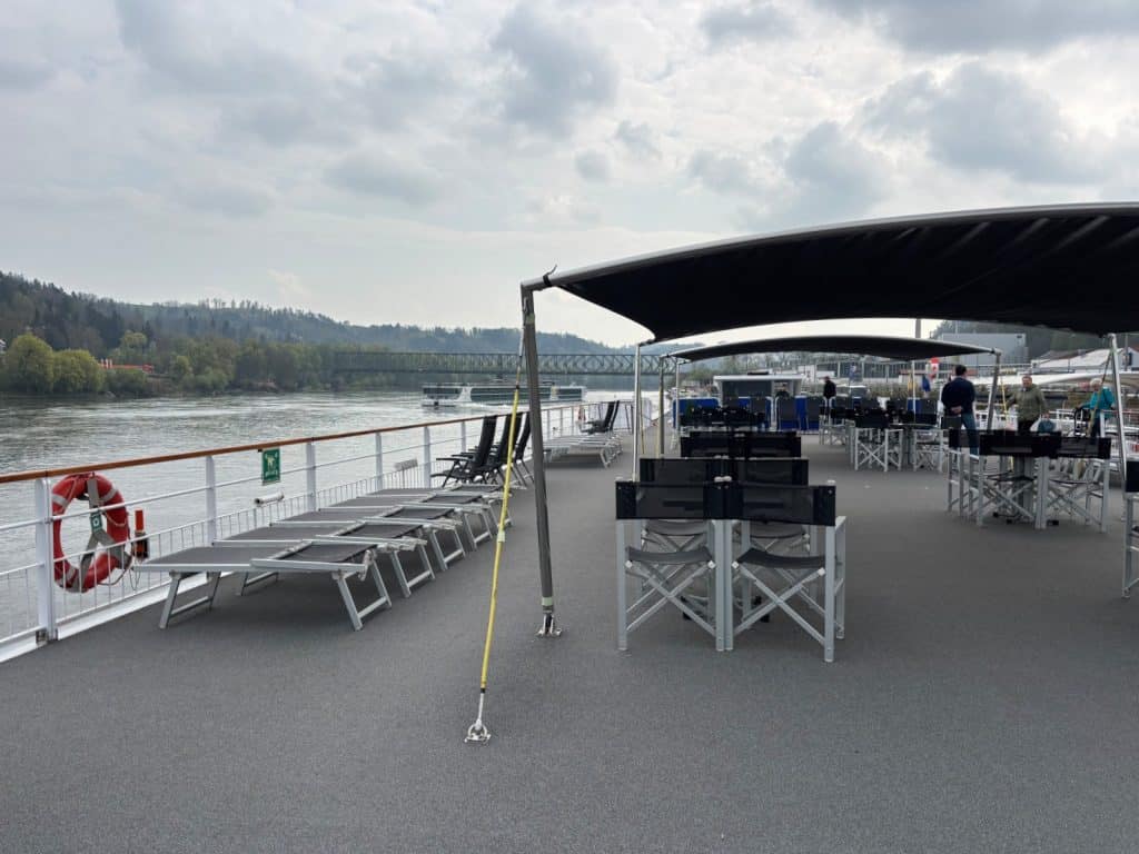 Das Sonnendeck des Flusskreuzfahrtschiffes MS Maxima mit grauen Liegestühlen, schwarzen Regiestühlen unter Sonnensegeln und Blick auf die Donau sowie eine Brücke im Hintergrund.