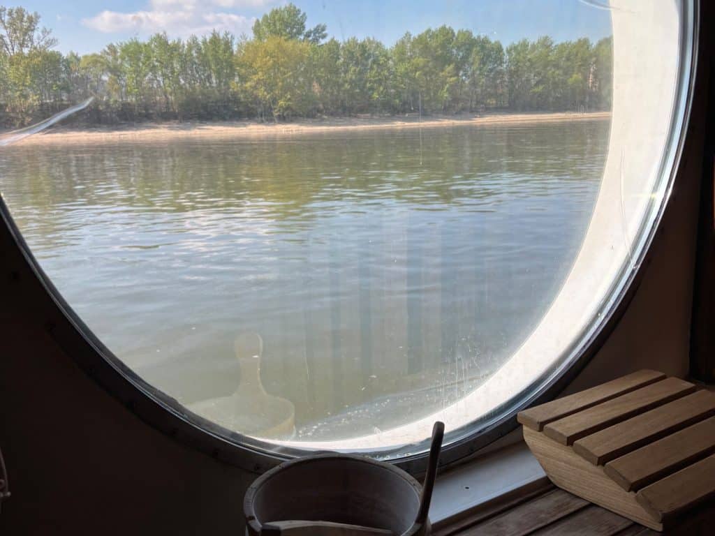 Blick aus einem runden Saunafenster der Maxima von nicko cruises auf den Fluss und ein bewaldetes Ufer, mit einem hölzernen Aufgusseimer und einer Kopfstütze im Vordergrund.