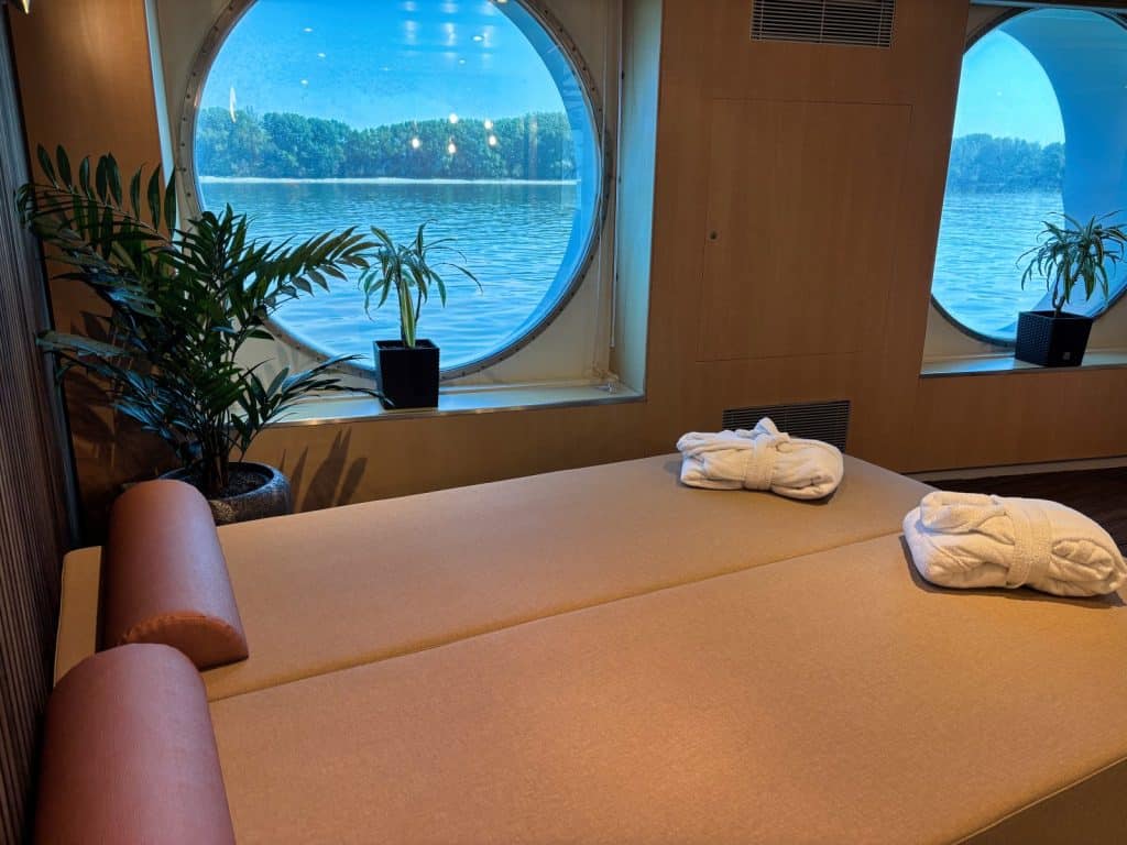 Zwei beigefarbene Wellnessliegen mit weißen Bademänteln im Spa-Bereich der Maxima von nicko cruises bieten durch große Bullaugenfenster einen direkten Blick auf das Wasser und die bewaldete Uferlandschaft.