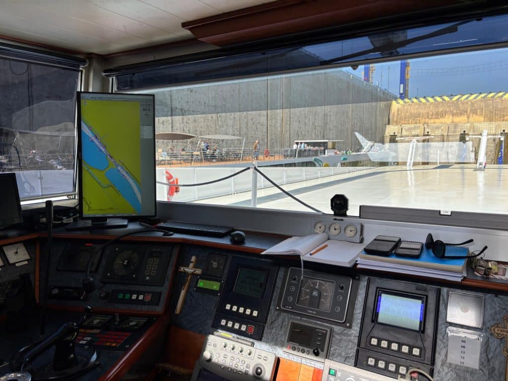 Blick aus dem Steuerhaus der Maxima von nicko cruises auf die Instrumententafel mit Navigationsmonitoren und das Außendeck, während das Schiff eine Schleuse passiert.