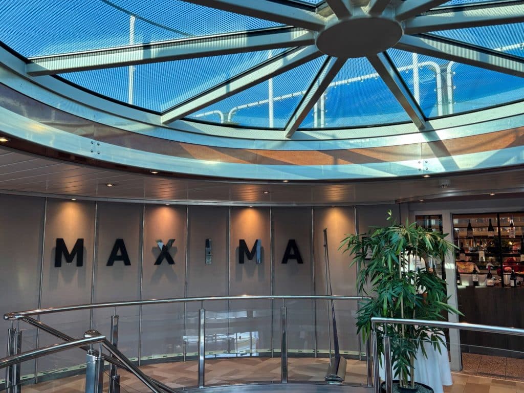 Innenansicht des Kreuzfahrtschiffs mit dem schwarzen Schriftzug „MAXIMA“ an einer geschwungenen Wand unter einer großen, runden Glaskuppel. Ein Treppenaufgang mit Edelstahlgeländer und eine große Grünpflanze befinden sich im Vordergrund.