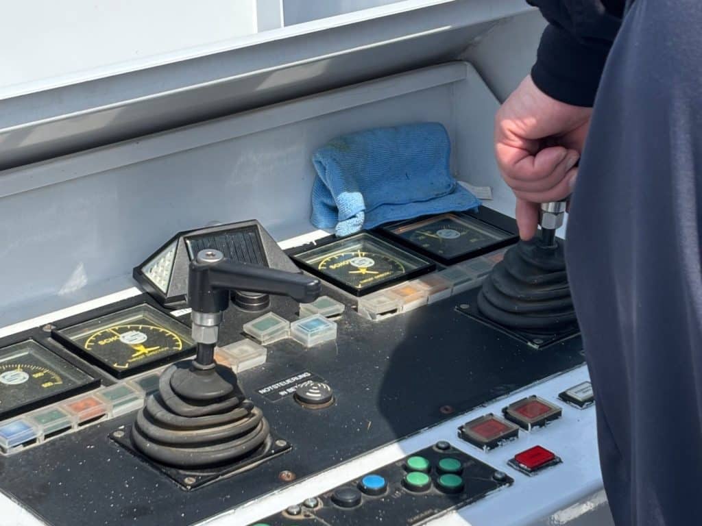 Nahaufnahme der Steuerkonsole auf der Maxima nicko cruises, bei der die Hand des Kapitäns einen Joystick bedient, während das Schiff aus einer Schleuse steuert. Das Bedienfeld zeigt verschiedene analoge Anzeigen, Hebel und farbige Knöpfe zur Navigation des Schiffes.