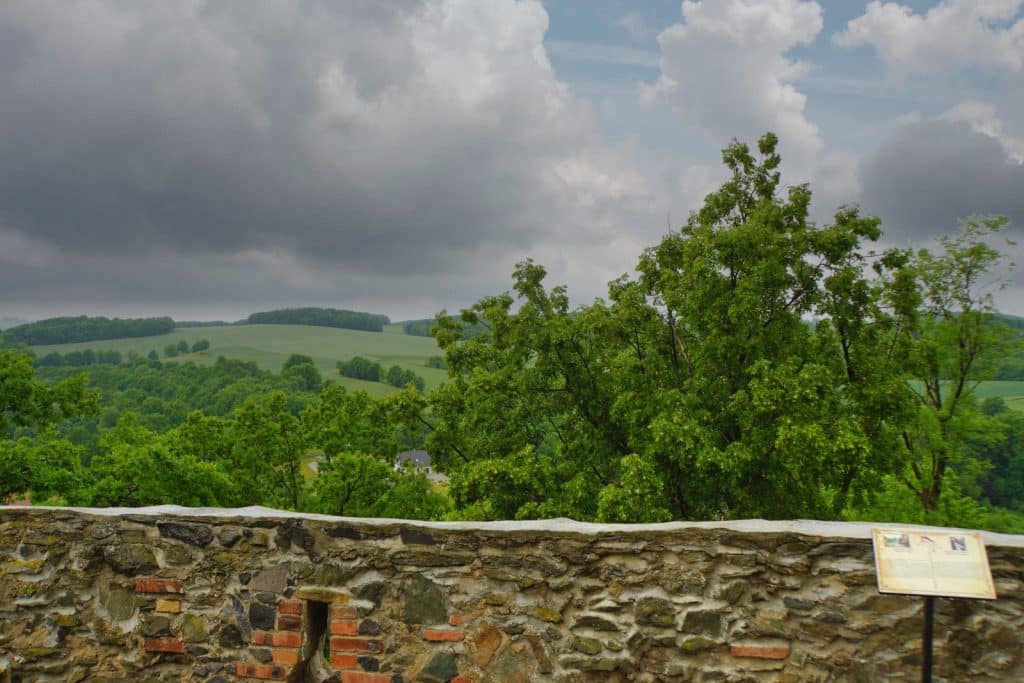 Blick von der Bolkoburg in Niederschlesien über eine steinerne Burgmauer auf eine hügelige, grün bewaldete Landschaft unter einem bewölkten Himmel.