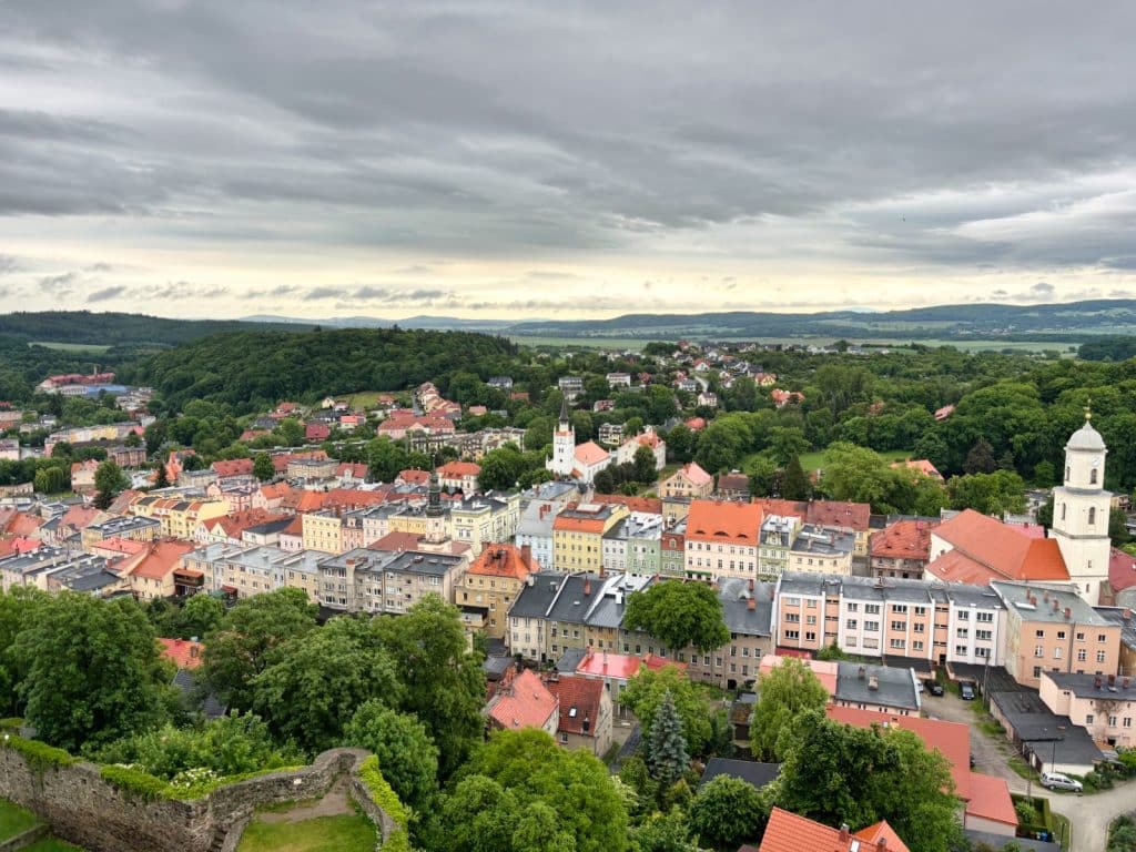 Blick von der Bolkoburg auf das Stadtzentrum von Bolków in Niederschlesien mit seinen bunten Häuserfassaden, Kirchtürmen und den umliegenden grünen Hügeln unter einem bewölkten Himmel.