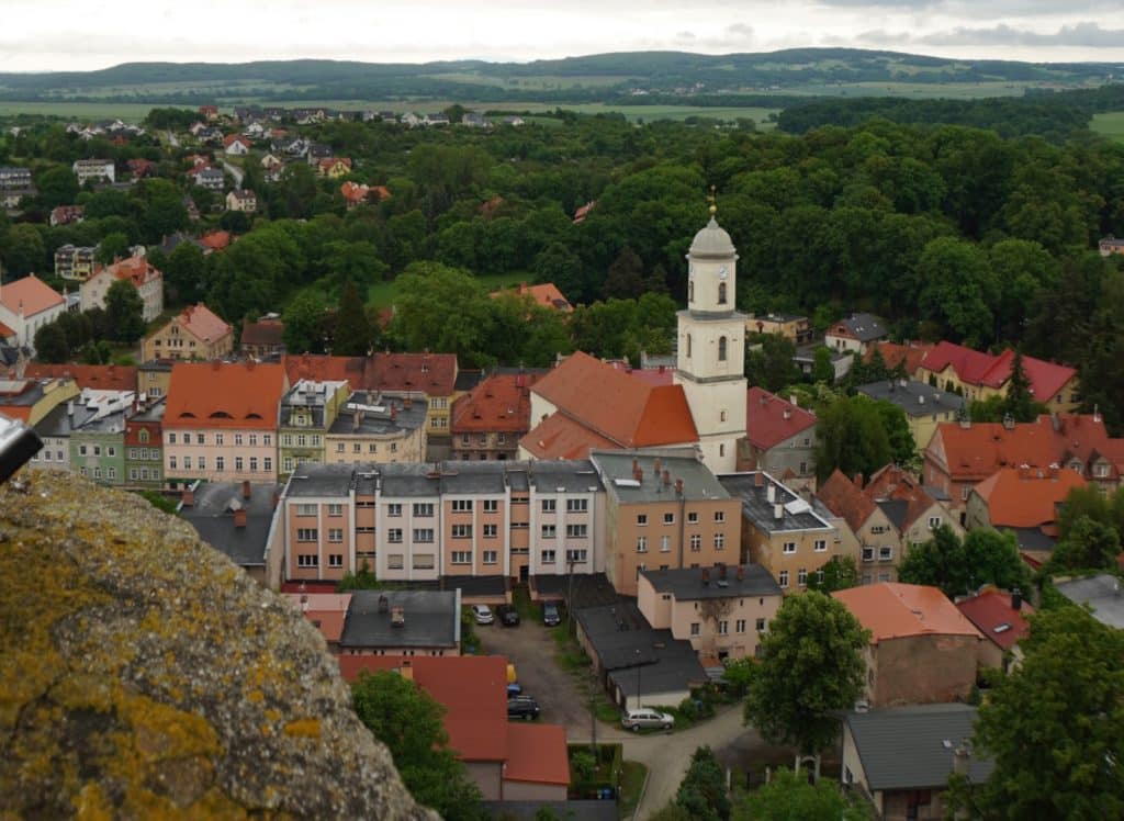Blick von der Bolkoburg auf das niederschlesische Dorf Bolków mit seinen eng beieinander liegenden Häusern mit roten Ziegeldächern und einem zentralen weißen Kirchturm vor einer bewaldeten Hügellandschaft.