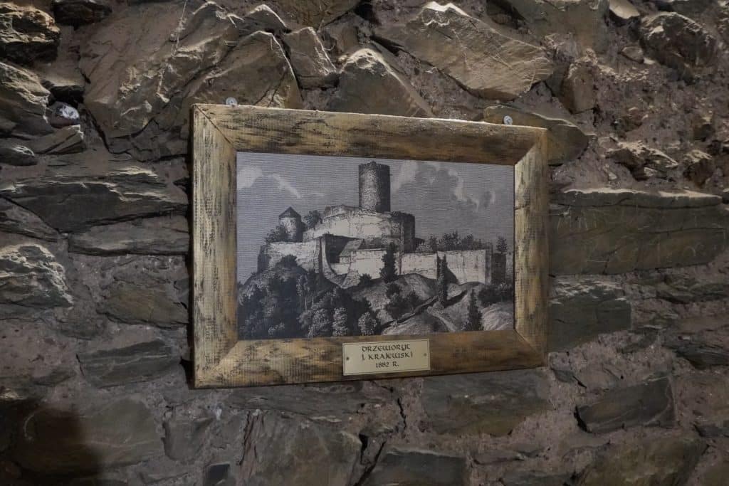 Ein gerahmtes historisches Bild an einer rustikalen Steinwand zeigt eine Schwarz-Weiß-Illustration der Bolkoburg in Niederschlesien aus dem Jahr 1882.