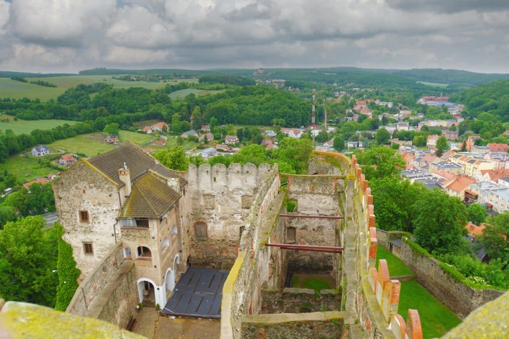 Blick von oben auf die Ruinen der Bolkoburg in Niederschlesien mit ihren erhaltenen Steinmauern und einem Wohngebäude, umgeben von einer grünen Hügellandschaft und einer angrenzenden Kleinstadt unter bewölktem Himmel.