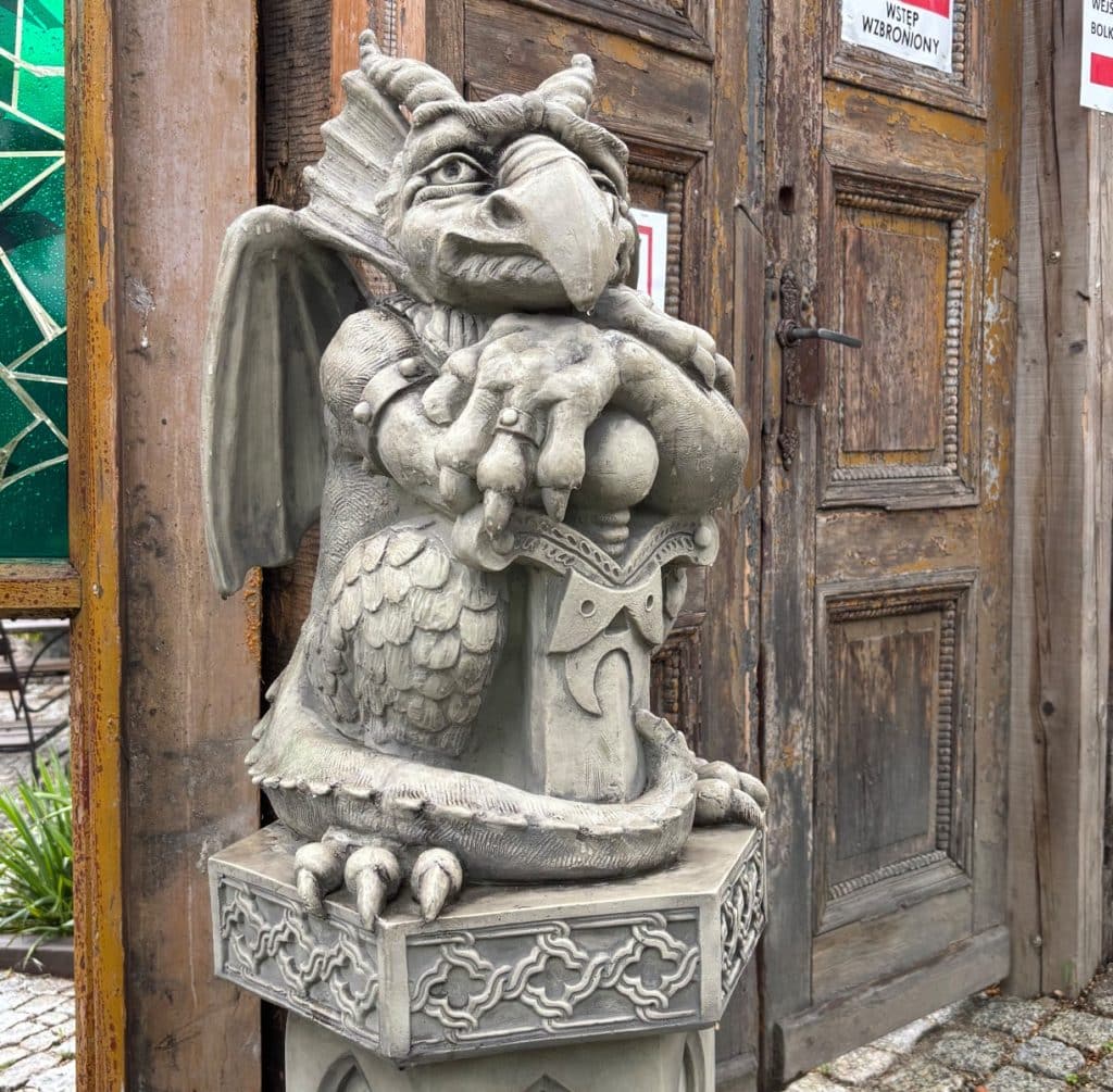 Eine steinerne Skulptur eines Torwächters in Form eines geflügelten Drachenwesens mit großem Schnabel bewacht den Eingang zur Bolkoburg in Niederschlesien. Die Figur sitzt auf einem verzierten Sockel vor einer alten, rustikalen Holztür und stützt ihre Klauen auf ein Schwert.