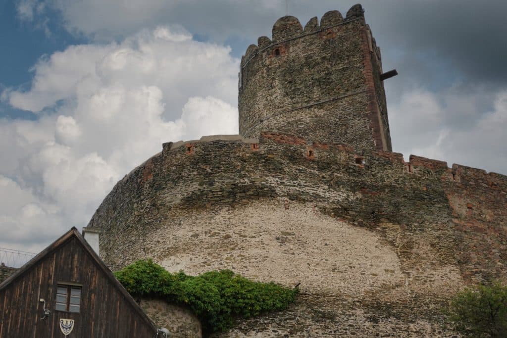 Der massive, runde Bergfried der Bolkoburg in Niederschlesien ragt hinter einer hohen, steinernen Wehrmauer unter einem bewölkten Himmel empor. Im Vordergrund ist das Giebeldach eines rustikalen Holzhauses mit einem kleinen Wappen zu sehen.